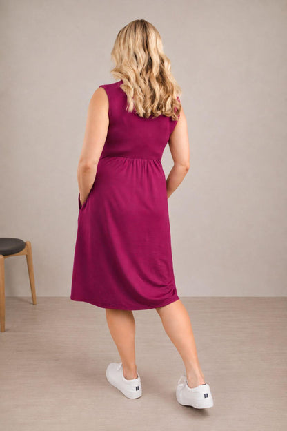 A woman wearing a sleeveless magenta midi dress with side pockets, shown from the back. She is standing in a minimalist room with a beige wall and light wood floor, wearing white sneakers.