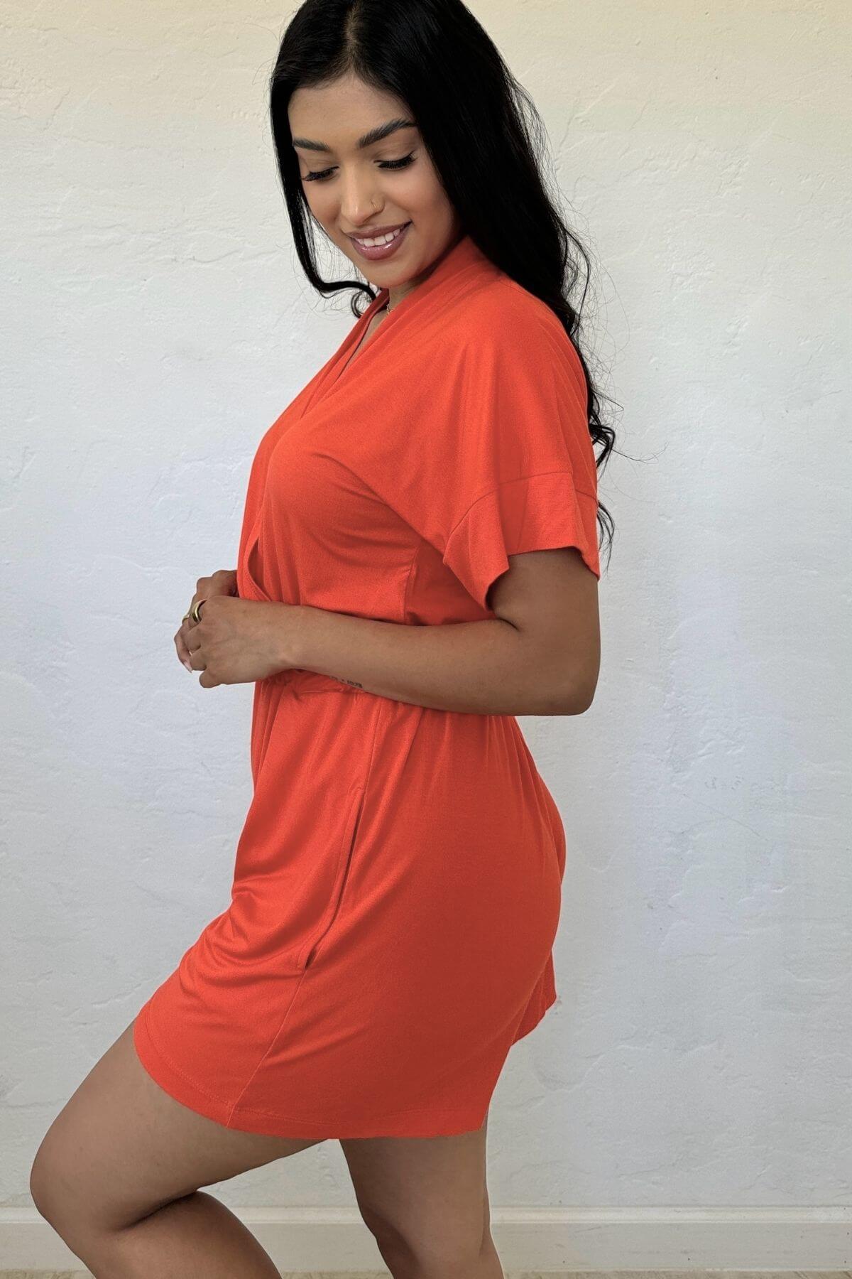 A woman modeling a bright orange short-sleeve mini dress against a textured off-white wall.