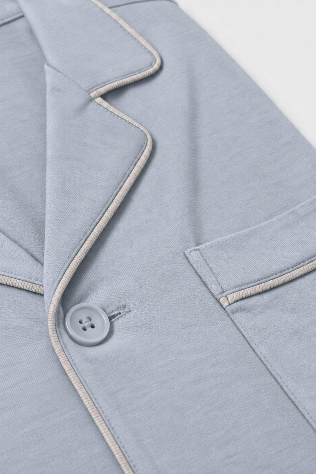 Close-up of a light blue-gray pajama top or long-sleeve top showing the collar, button closure, and a pocket with contrasting light beige piping.