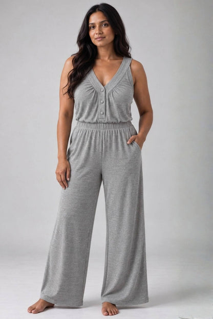 A woman modeling a light gray sleeveless jumpsuit with a V-neck, button-front bodice, elastic waist, and wide legs, photographed against a plain light gray studio background.
