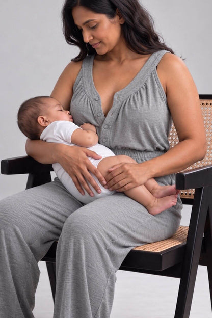 A woman wearing a gray sleeveless baby nursing top and matching pants sits on a black chair with a woven seat, holding a sleeping baby dressed in a white onesie.