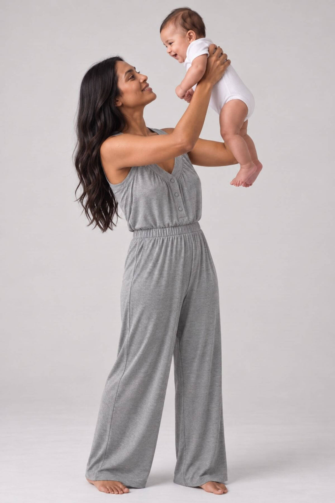 A woman in a heather gray sleeveless jumpsuit holds a baby wearing a white bodysuit against a plain light gray studio background.