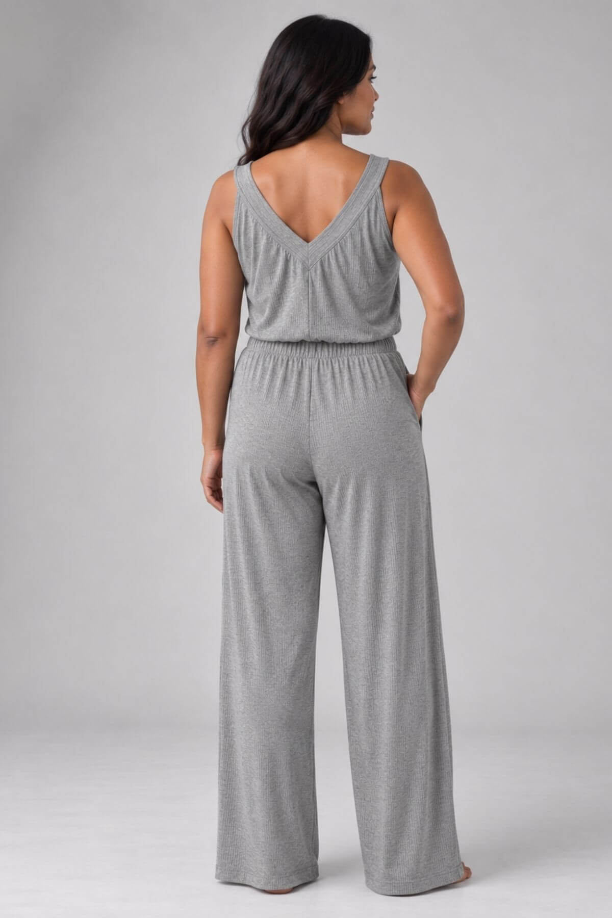 A woman wearing a heather gray sleeveless jumpsuit, shown from the back. The jumpsuit features a V-neckline and wide-leg pants, photographed against a neutral light gray studio background.