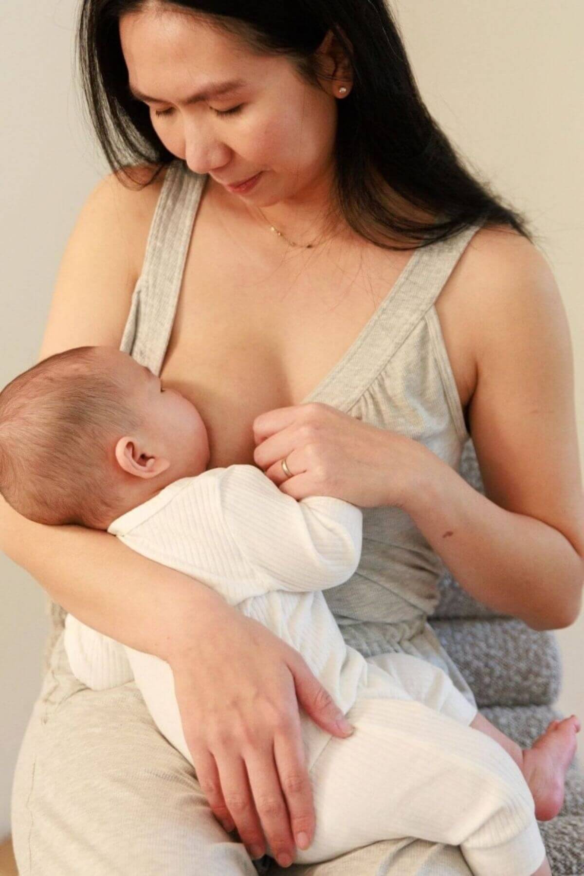 A woman in a light gray tank top and matching pants holds a baby dressed in a white ribbed onesie while breastfeeding. The photo is taken in a softly lit indoor setting with a neutral beige background.