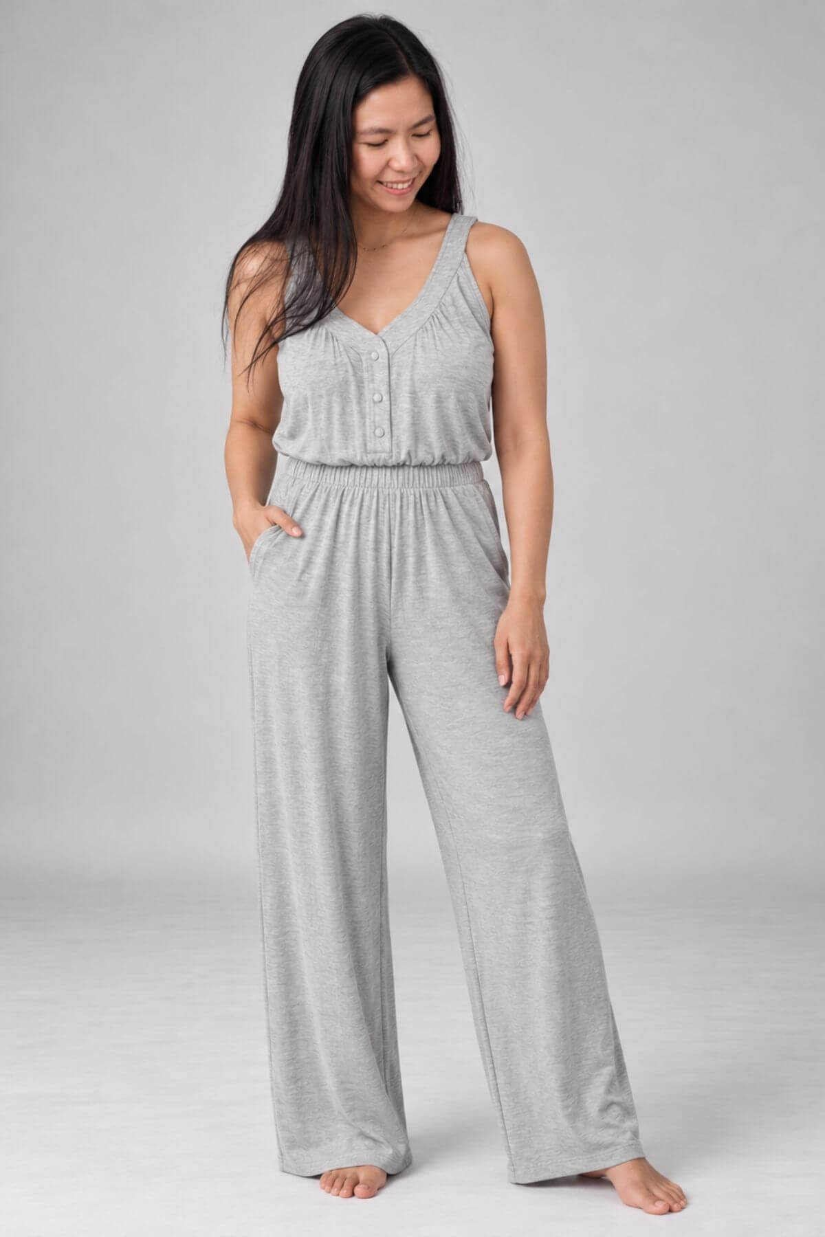 A woman modeling a light gray sleeveless jumpsuit with a buttoned bodice and wide-leg pants, standing against a neutral light gray studio background.