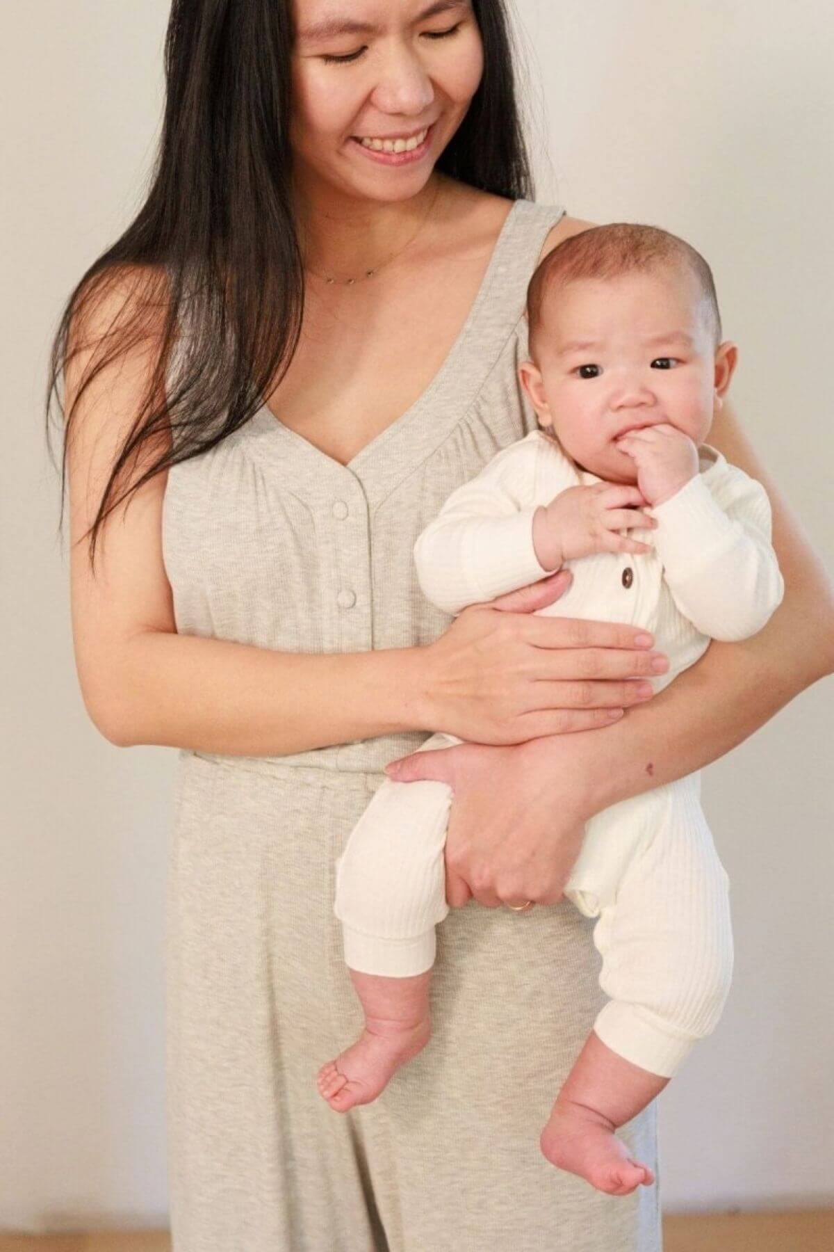 A smiling woman in a light beige sleeveless top holds a baby wearing a cream ribbed long-sleeve romper against a plain light beige background.