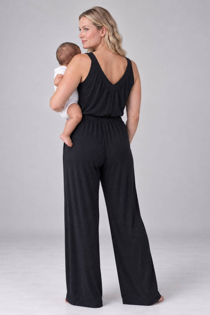A woman wearing a black sleeveless jumpsuit with wide legs and a backless V-neck, holding a baby against her chest, photographed against a light gray studio background.