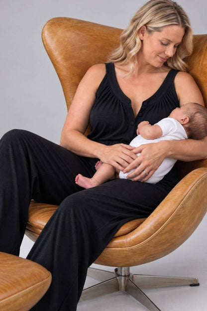 A woman in a black sleeveless top and black pants sits in a tan leather chair, holding a baby dressed in a white outfit. The background is a clean, neutral gray studio backdrop.