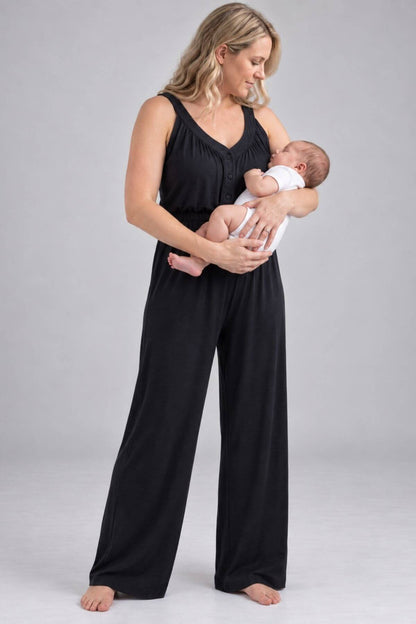 A woman wearing a black sleeveless jumpsuit with wide legs holds a sleeping newborn baby dressed in a white outfit, standing against a neutral light gray studio background.