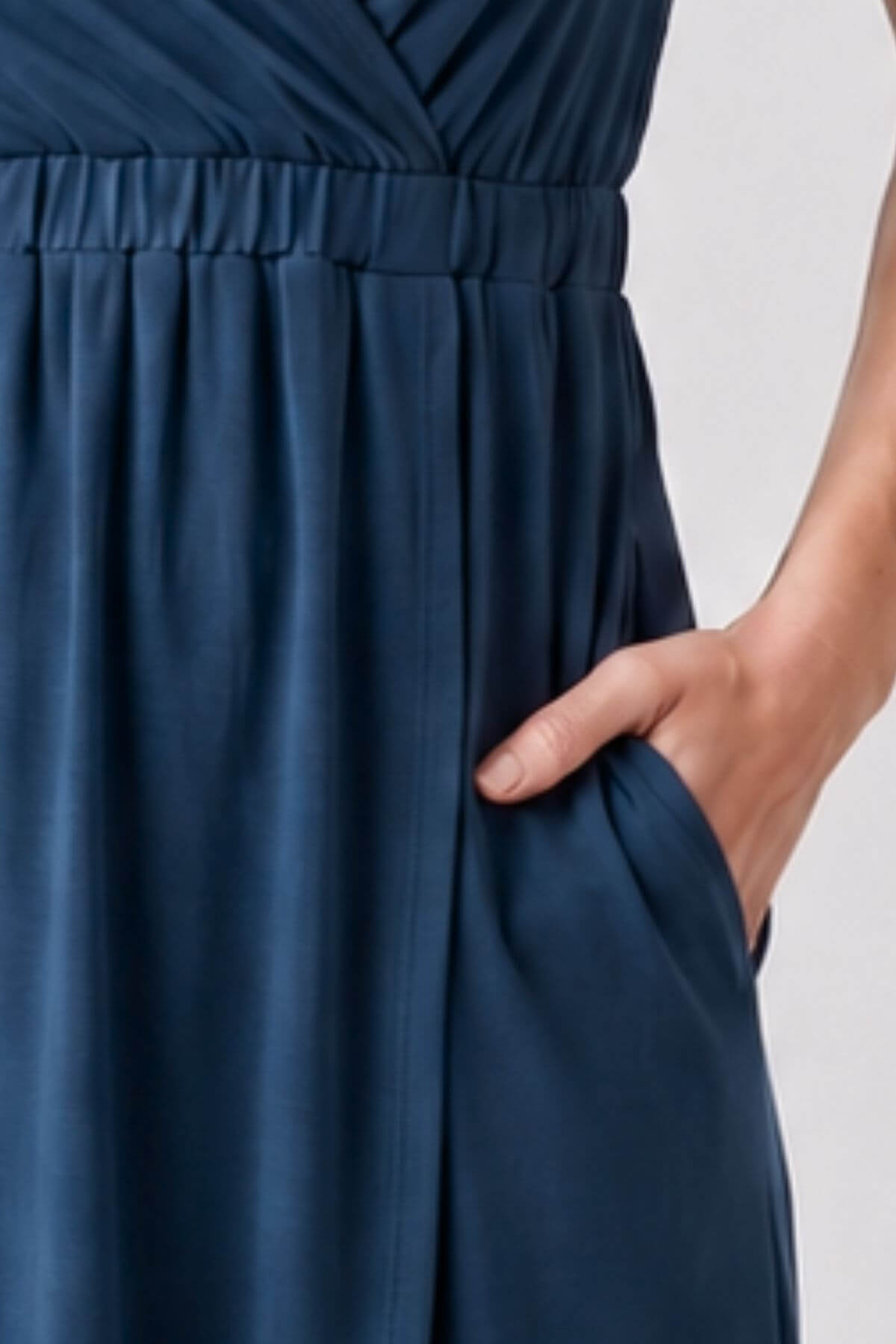 Close-up of a navy blue dress with a gathered elastic waistband and side pocket, shown on a model with one hand in the pocket against a light gray background.