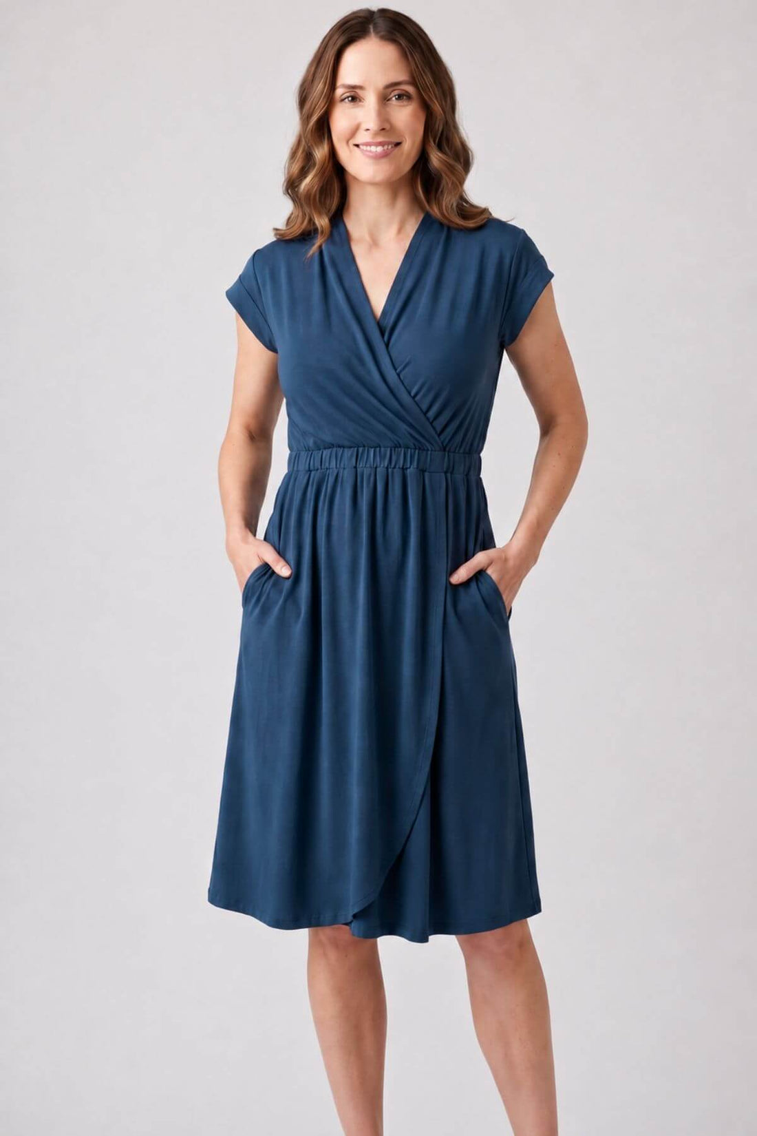 A woman modeling a navy blue wrap-style midi dress with cap sleeves, a V-neckline, an elastic waist, and side pockets, set against a plain light gray studio background..