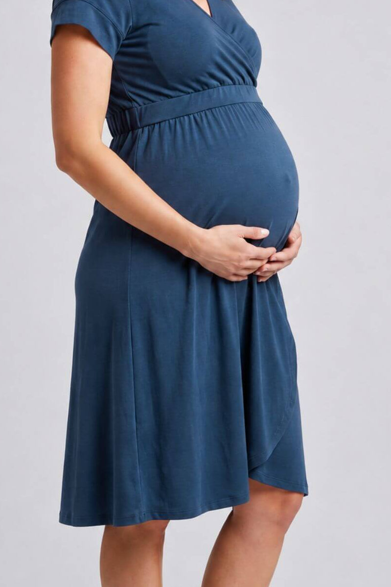 A pregnant woman wearing a slate blue short-sleeve maternity dress, standing against a light gray studio background..