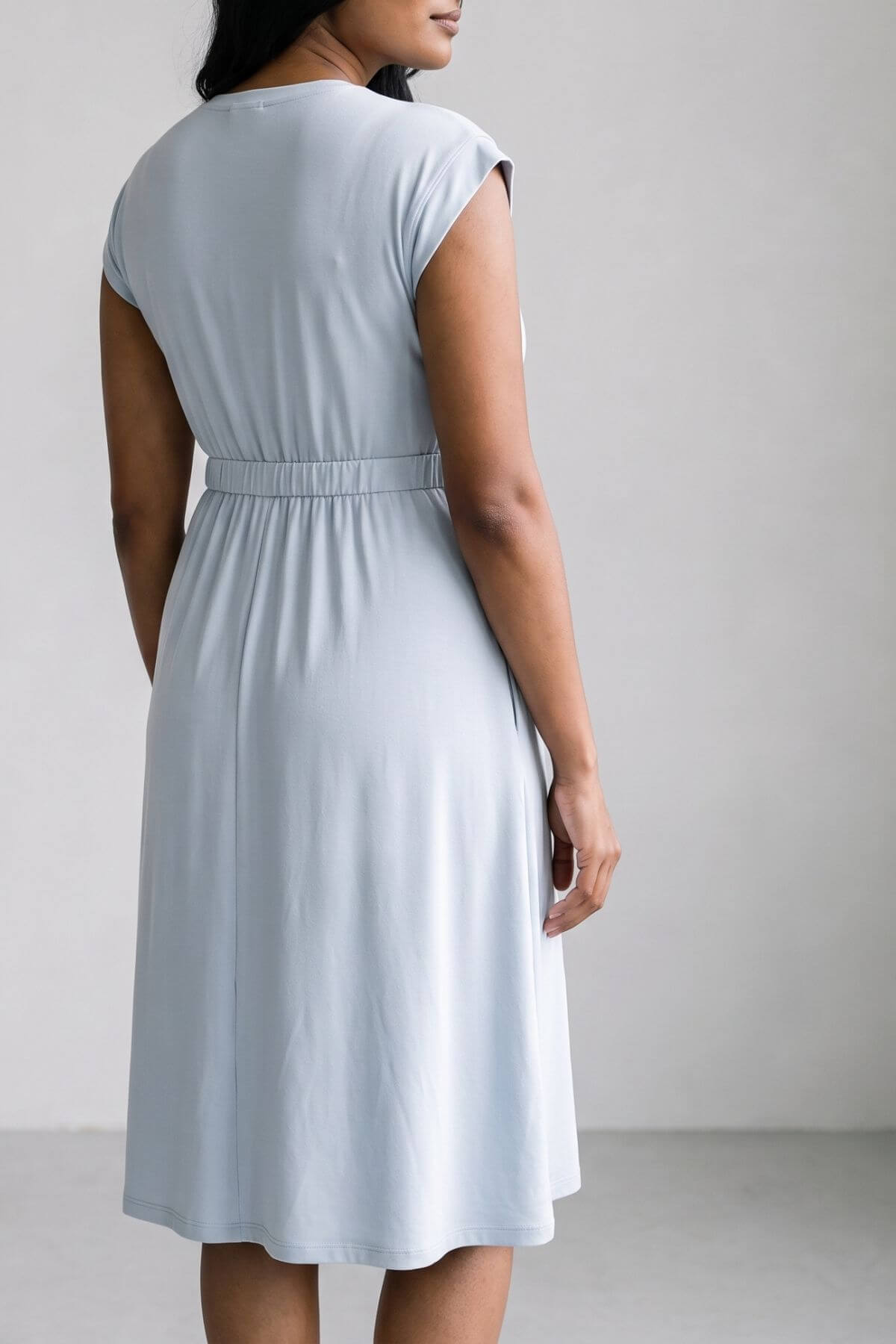 A woman wearing a light blue sleeveless dress with a matching waist tie, shown from the back against a neutral gray studio background.