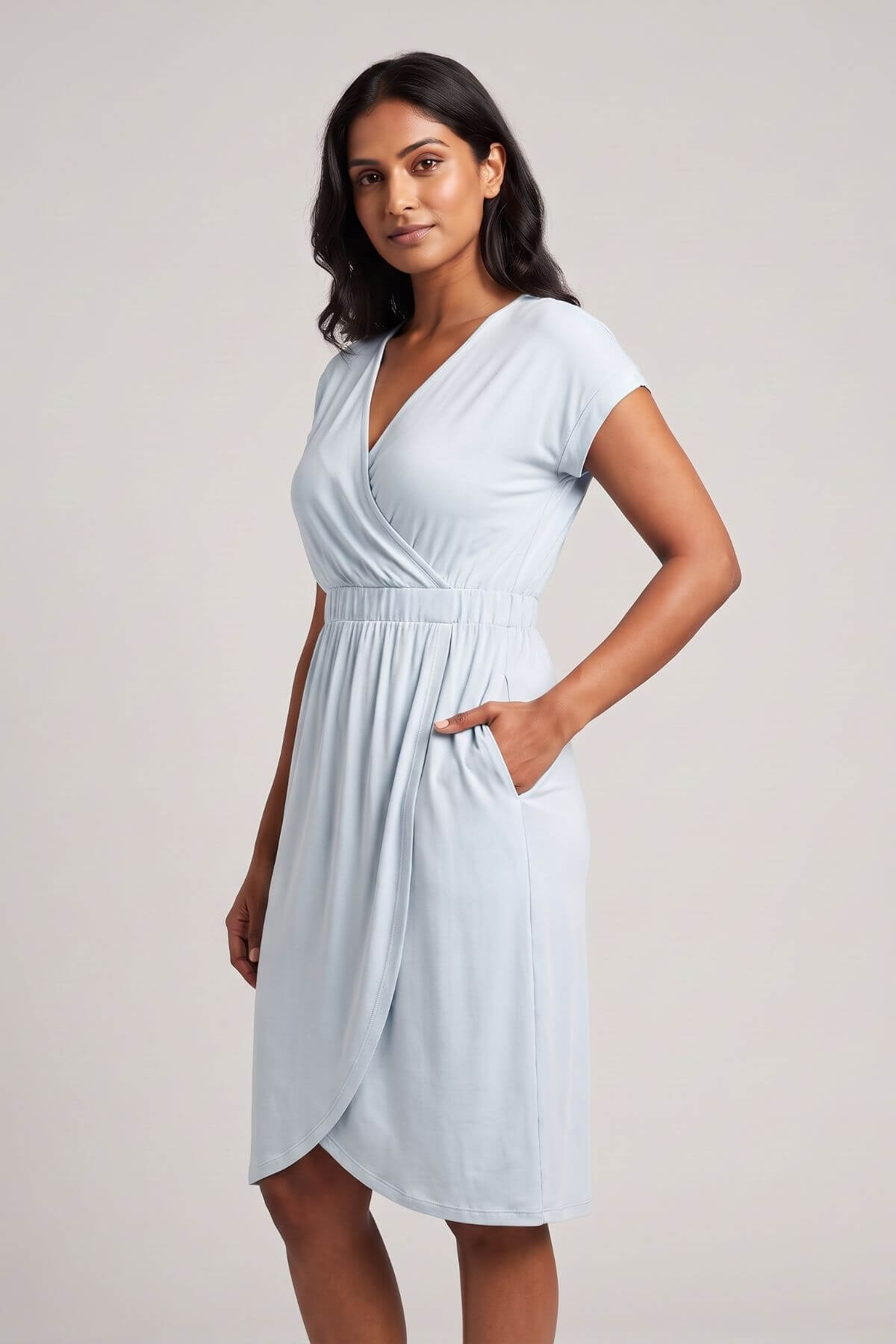 A woman modeling a light blue wrap-style midi dress with short sleeves, a V-neckline, and side pockets against a plain light gray studio background.