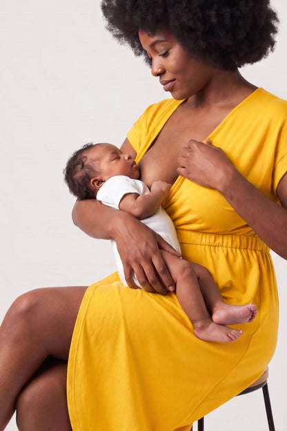 A woman in a bright yellow dress breastfeeding a baby wearing a white onesie, seated against a plain light gray studio background.