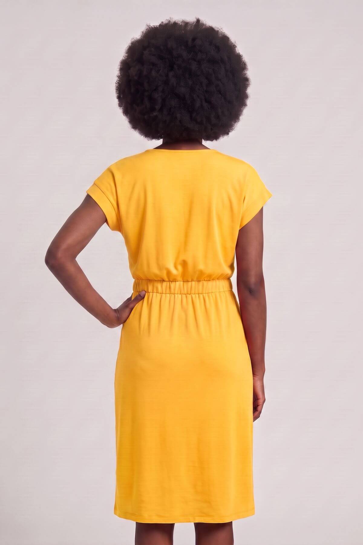A woman with a large dark afro is shown from the back wearing a bright yellow short-sleeve dress with a gathered elastic waist, set against a plain light gray studio background..