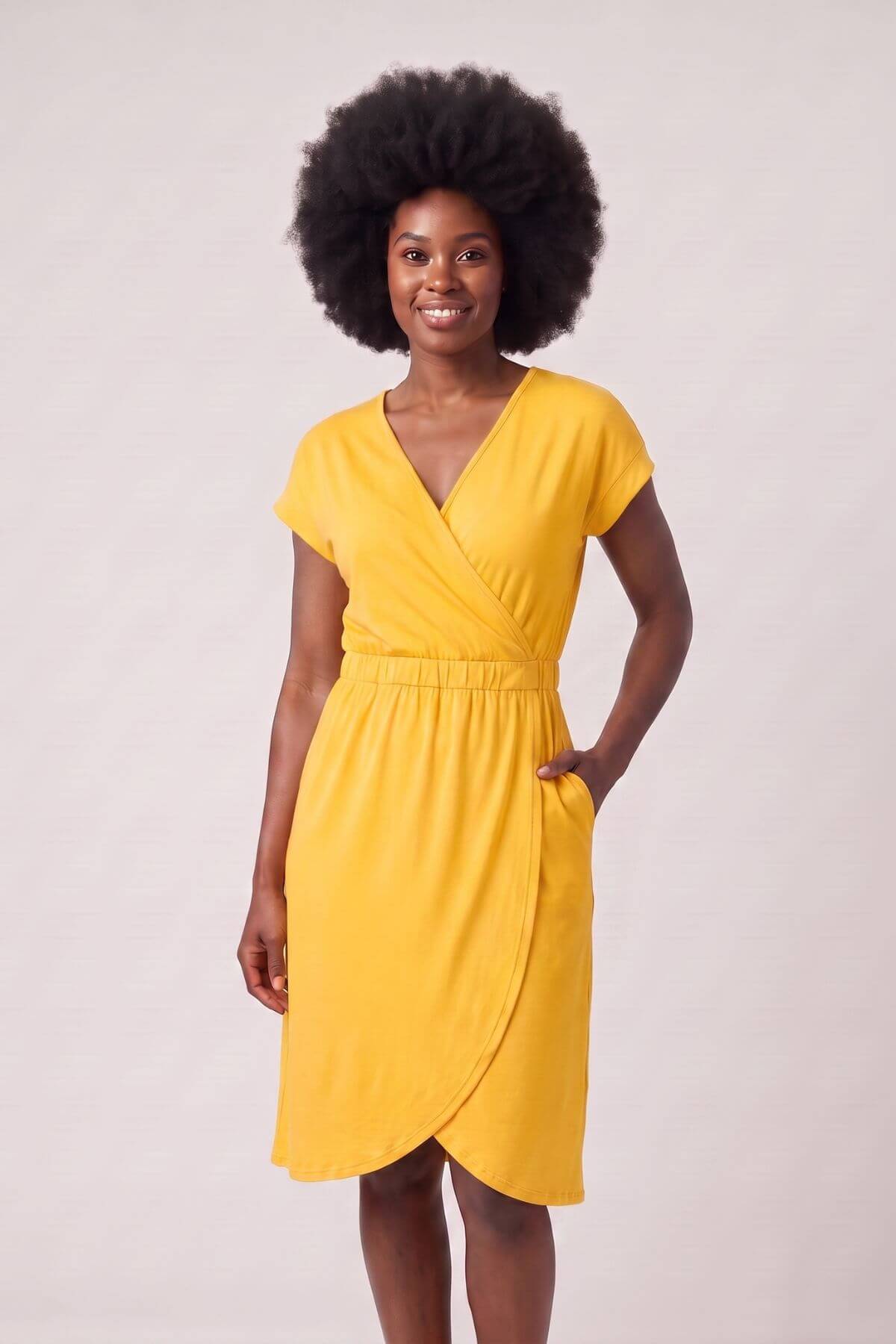 A woman with a large dark afro wearing a bright yellow wrap-style dress with short sleeves and a V-neckline, standing against a plain light beige studio background..