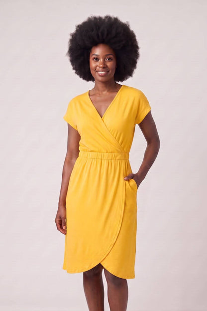 A woman with a large dark afro wearing a bright yellow wrap-style dress with short sleeves and a V-neckline, standing against a plain light beige studio background..