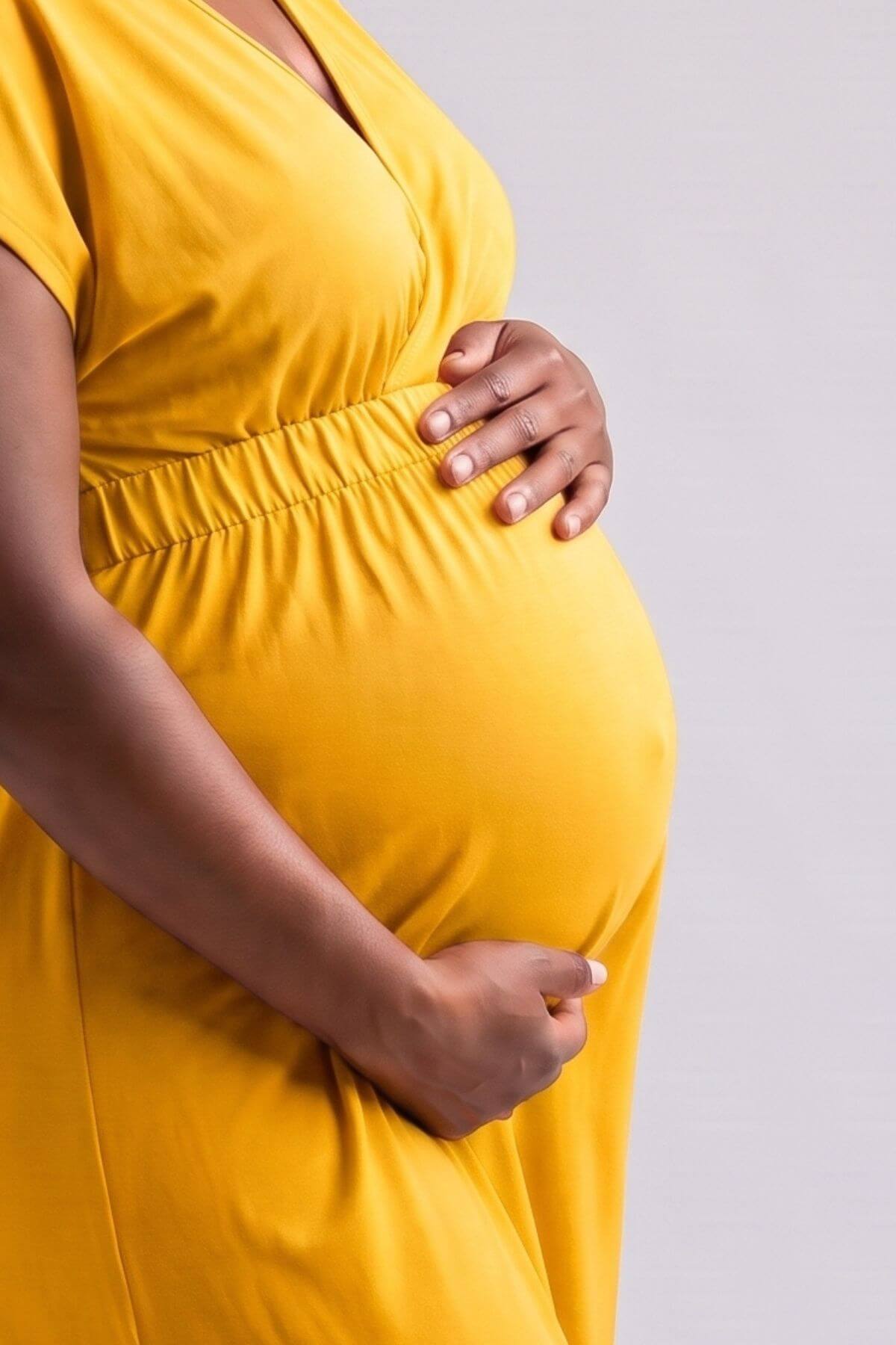 A close-up of a pregnant person wearing a bright yellow maternity dress, holding their belly against a plain light gray studio background..