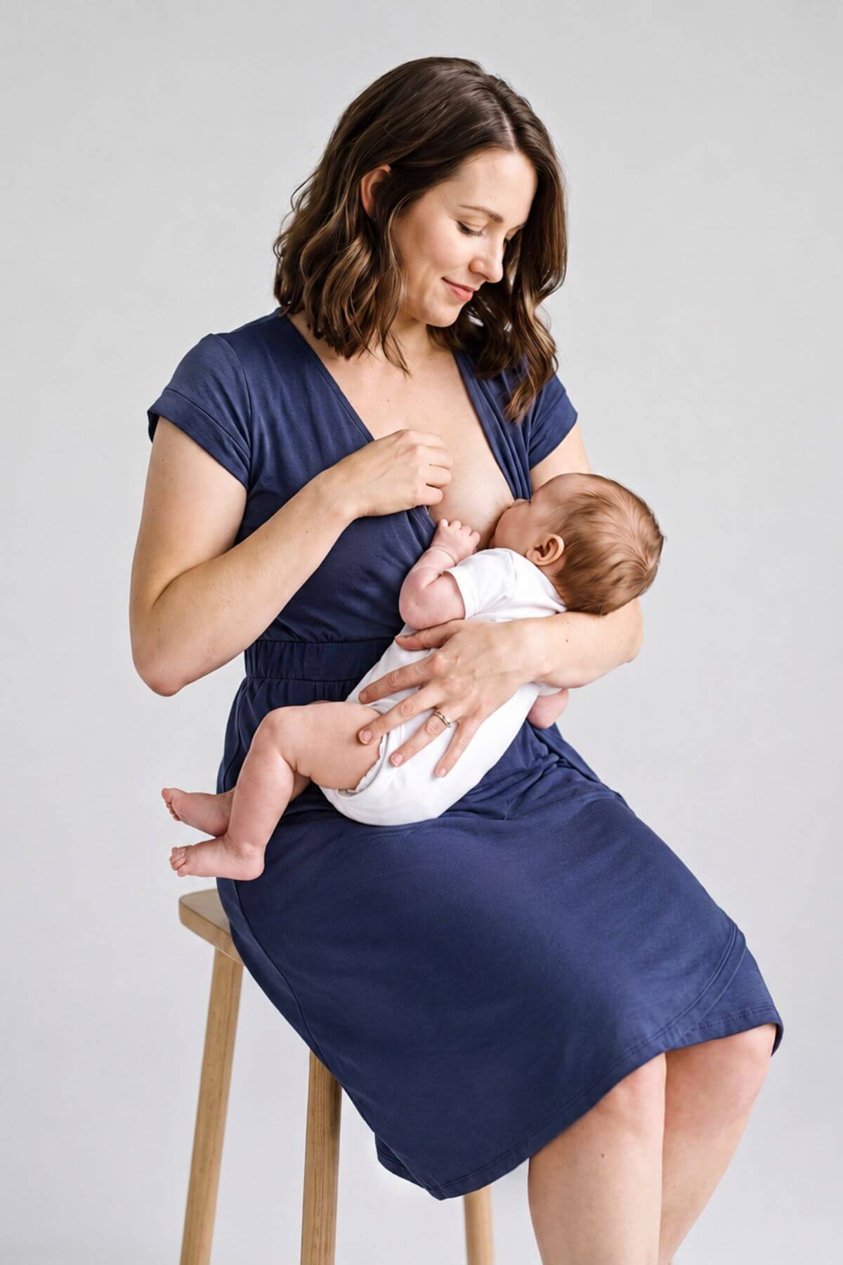 A woman wearing a navy blue maternity nursing dress sits on a wooden stool, breastfeeding a baby wrapped in a white swaddle. The photo is shot in a studio setting against a light gray background.