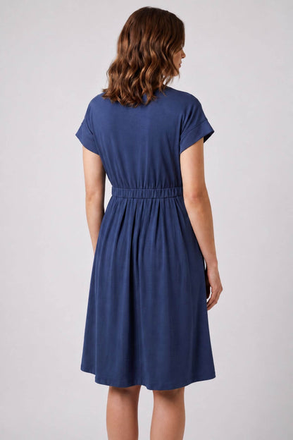 A woman viewed from the back wearing a navy blue short-sleeve dress with a gathered waist, set against a plain light gray studio background.