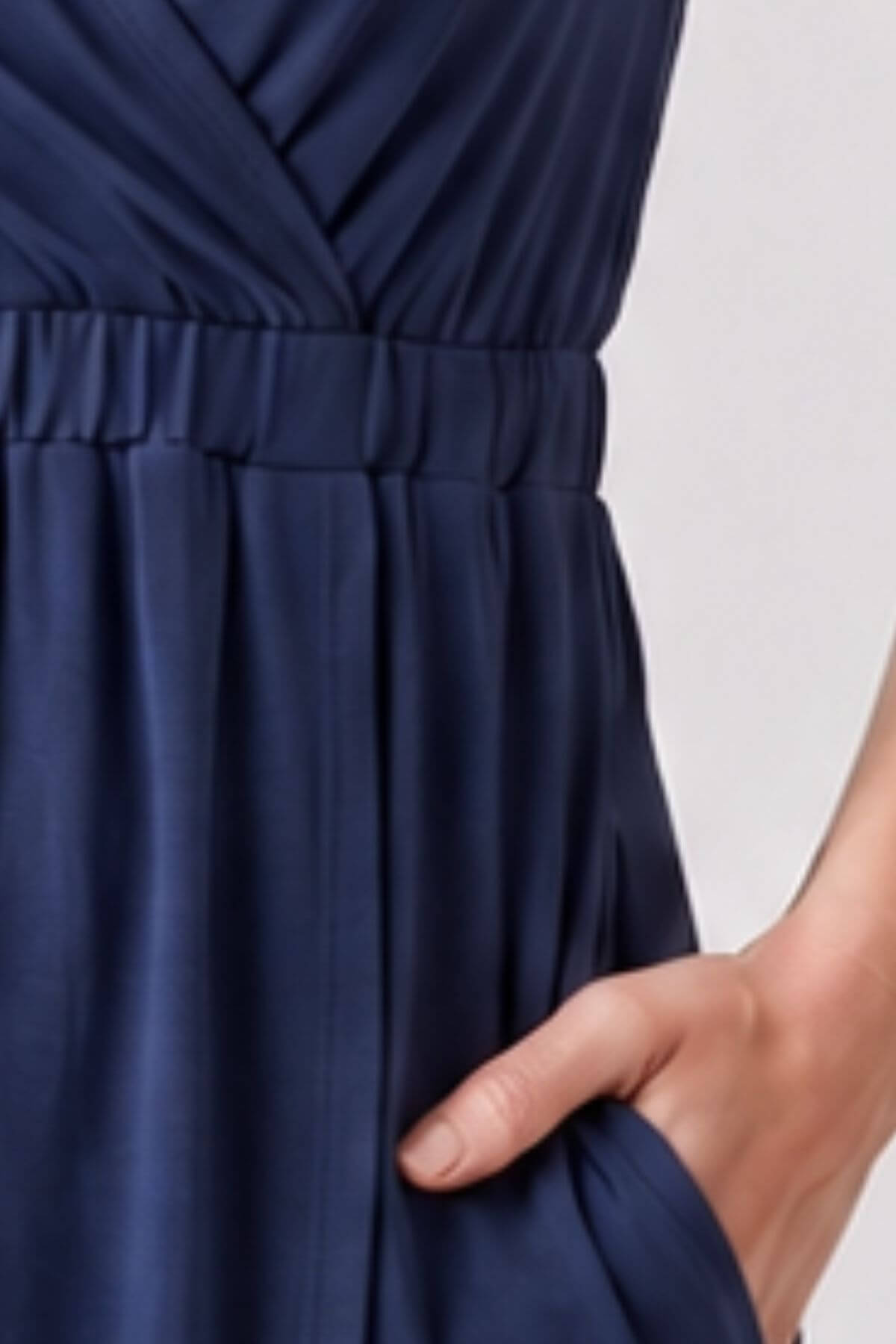 Close-up of a navy blue dress with a gathered waist and a hand tucked into a side pocket, against a light neutral background.