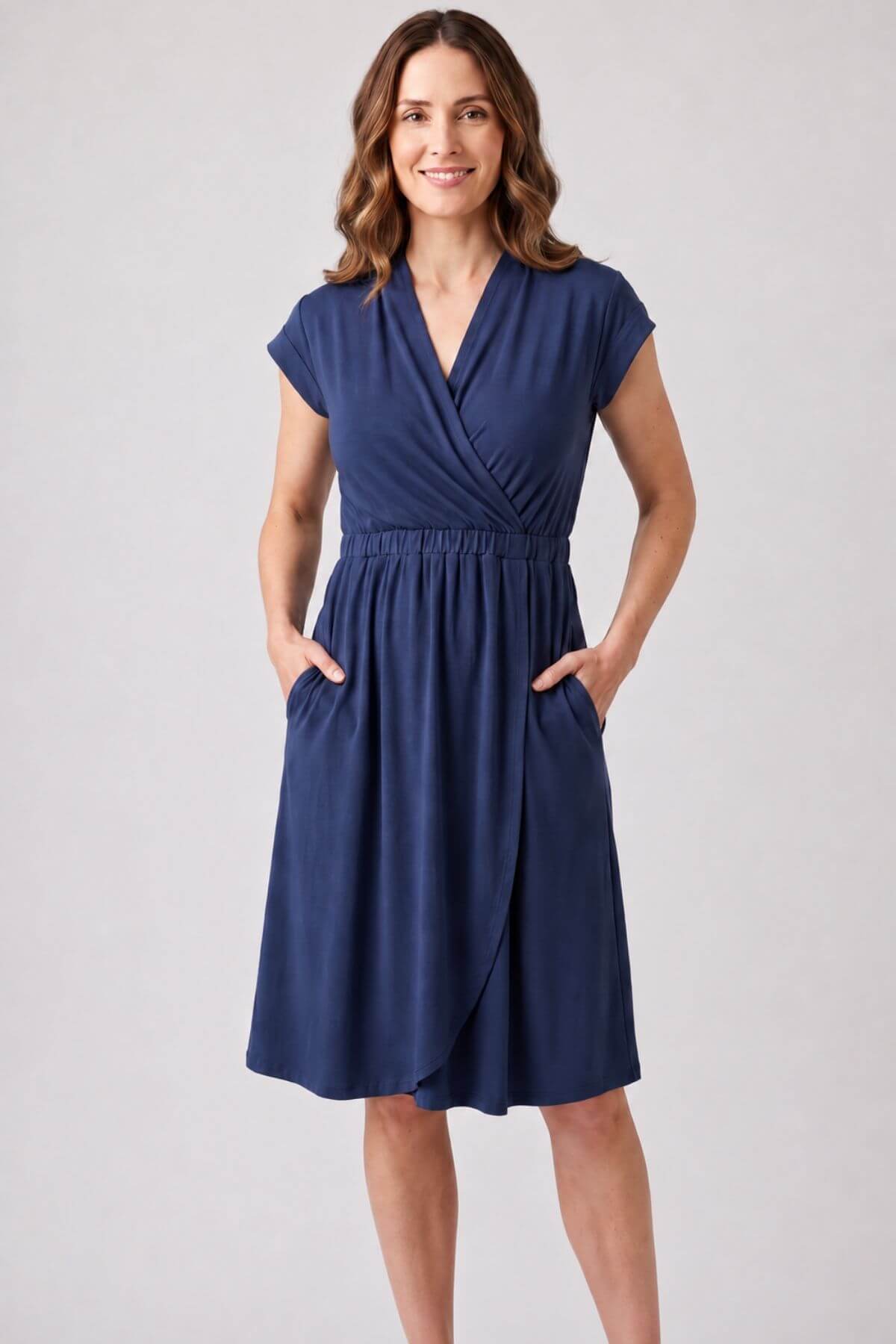 A woman modeling a navy blue wrap-style dress with cap sleeves, a V-neckline, an elastic waist, and side pockets, photographed against a plain light gray studio background.