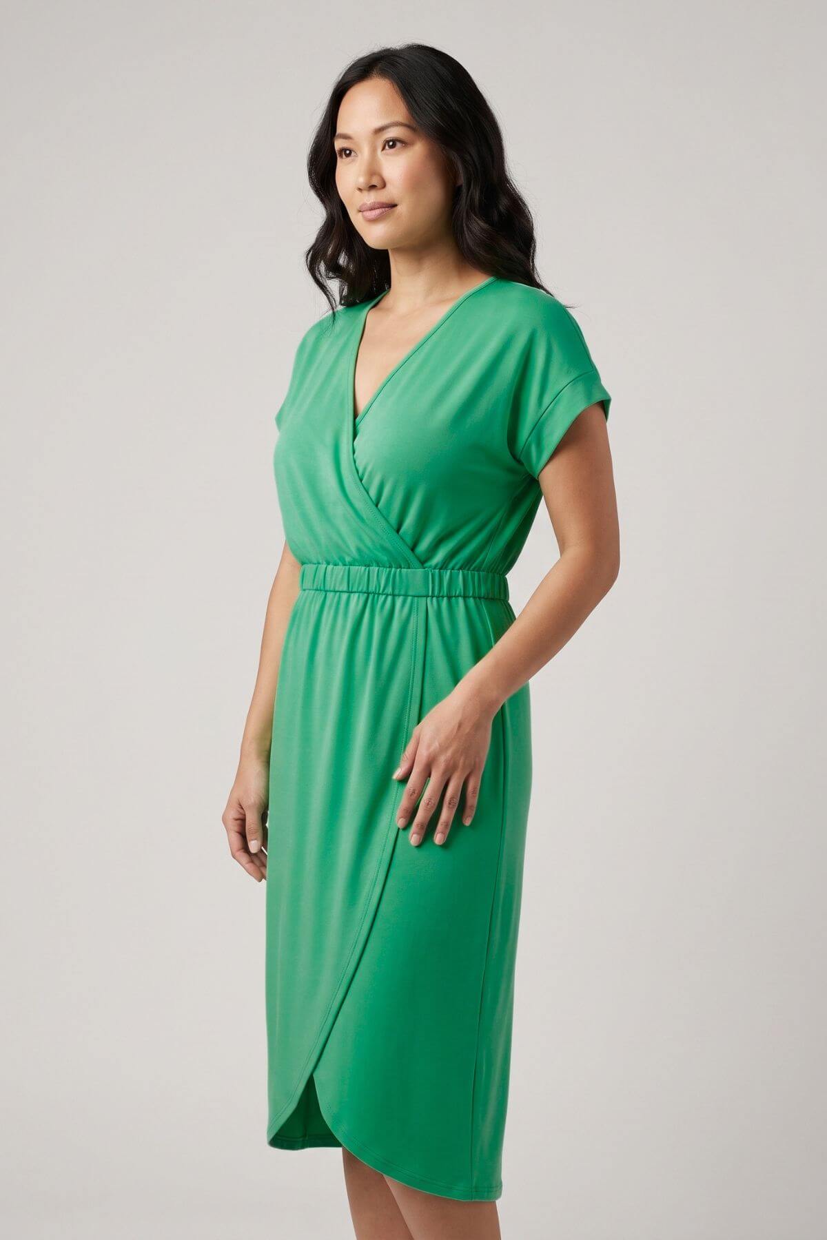 A woman modeling a bright green wrap-style midi dress with short sleeves and a V-neckline, photographed against a plain light gray studio background.