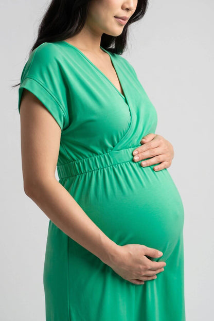 A pregnant woman wearing a bright green short-sleeve wrap dress, holding her baby bump against a light gray studio background.