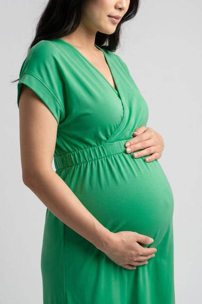 A pregnant woman wearing a bright green short-sleeve maternity dress with a V-neck and gathered elastic waist, standing against a light gray studio background.