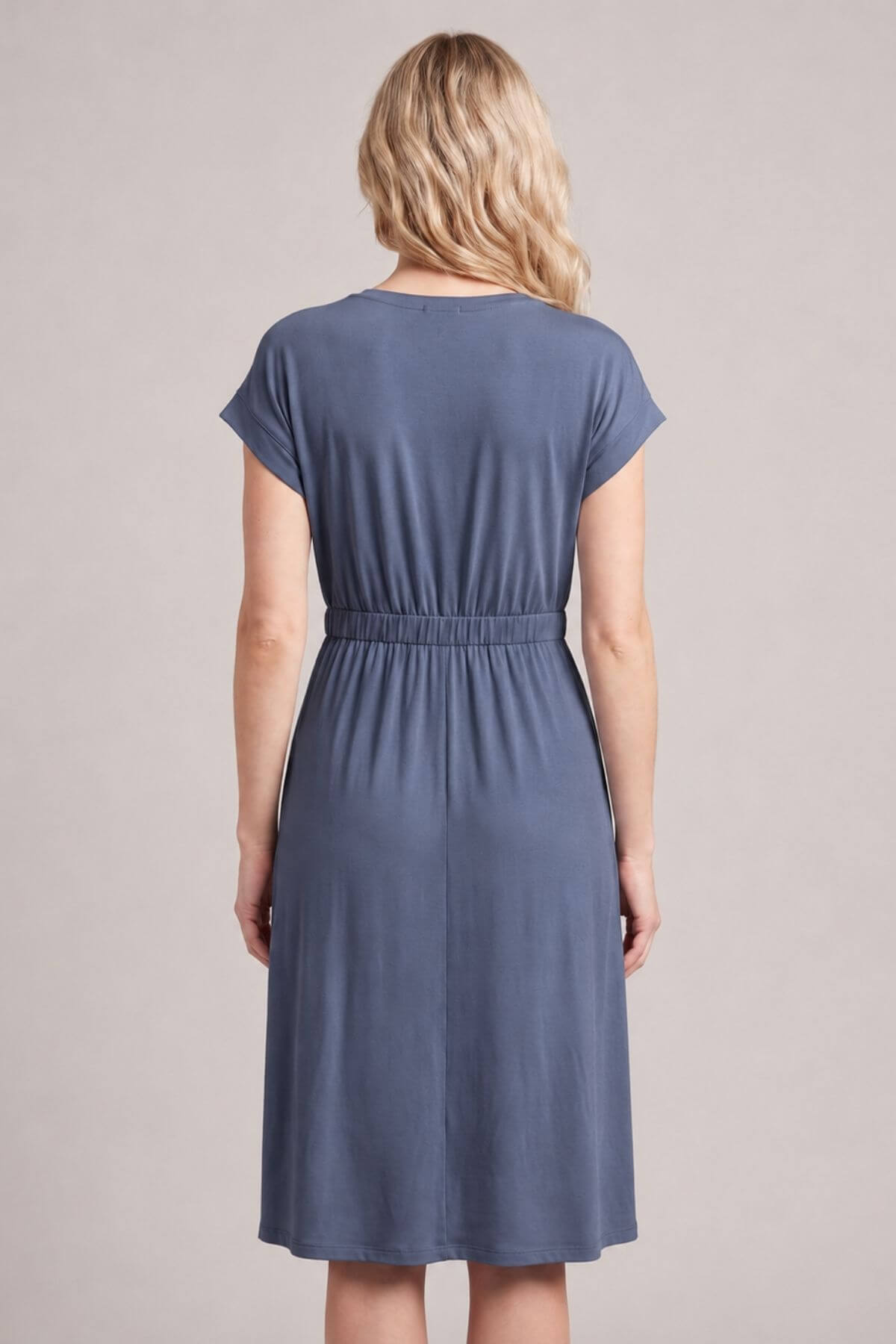 A woman viewed from the back wearing a slate blue short-sleeve dress with a gathered elastic waist, set against a plain light gray studio background.