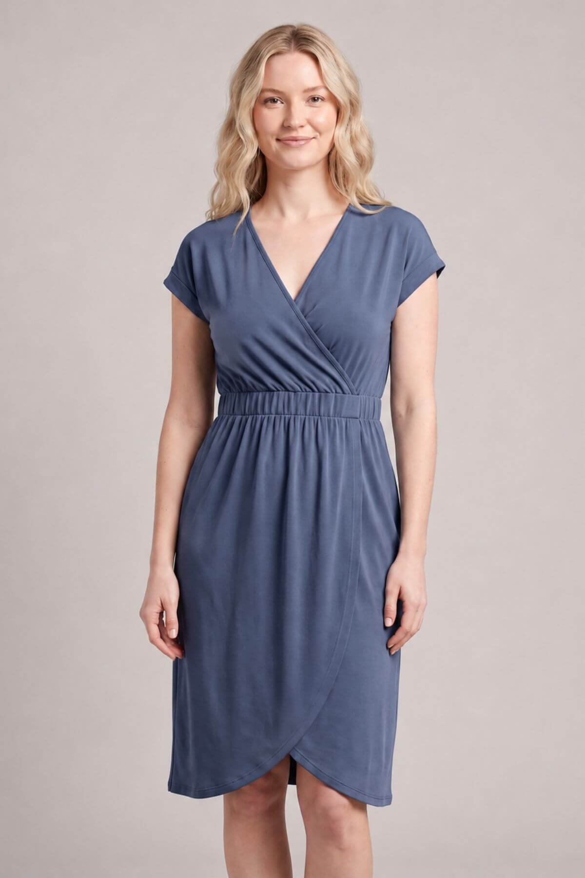 A woman modeling a slate blue wrap-style dress with short cap sleeves, a V-neckline, and a gathered waist, photographed against a plain light gray studio background.