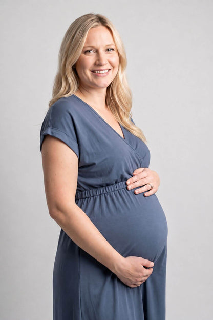 A smiling pregnant woman wearing a slate blue maternity dress, standing against a neutral light gray studio background.