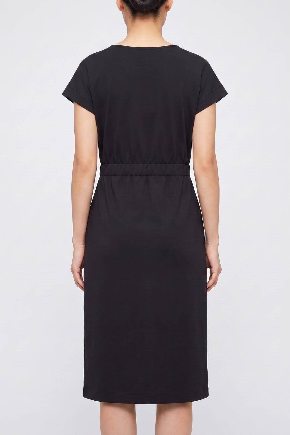 A woman viewed from the back wearing a black short-sleeve midi dress with a defined waist seam, set against a plain light gray studio background.