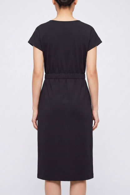 A woman viewed from the back wearing a black short-sleeve midi dress with a defined waist seam, set against a plain light gray studio background.