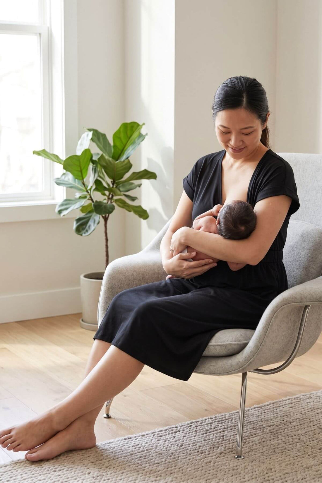 A woman in a black dress sits in a light gray armchair, holding a baby. The scene is set in a bright, modern room with a potted fiddle-leaf fig plant, light wood floors, and a textured rug.