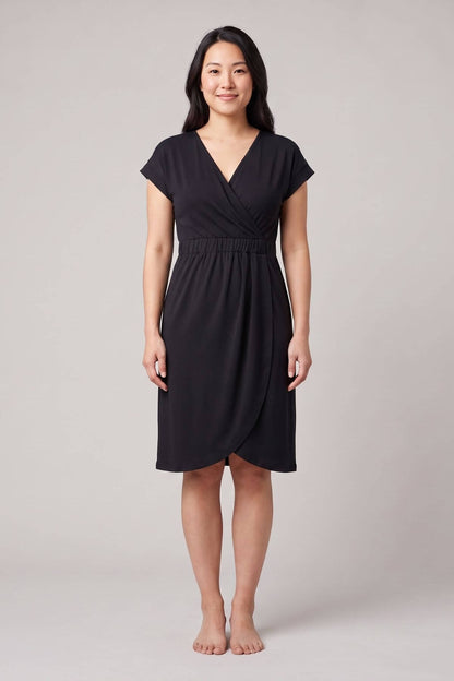 A woman modeling a black short-sleeve wrap-style dress with a V-neckline and gathered waist, standing against a plain light gray studio background.