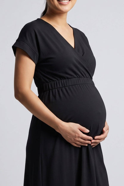 A pregnant woman wearing a black short-sleeve wrap-style maternity dress, holding her baby bump against a light gray studio background.