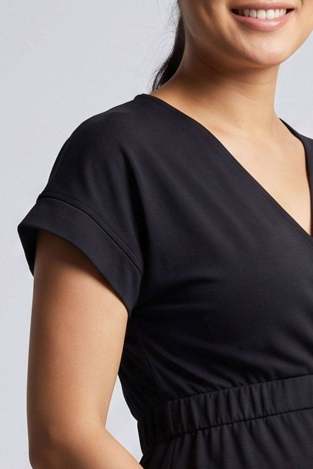 Close-up of a woman wearing a black short-sleeve V-neck dress or top with a gathered waist, shown against a light gray studio background.