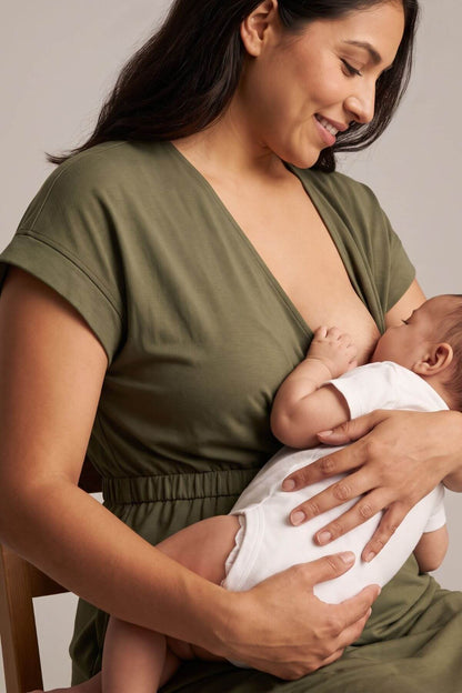 A woman in an olive green short-sleeve V-neck dress is breastfeeding a baby wearing a white onesie while seated on a wooden chair against a neutral beige background.