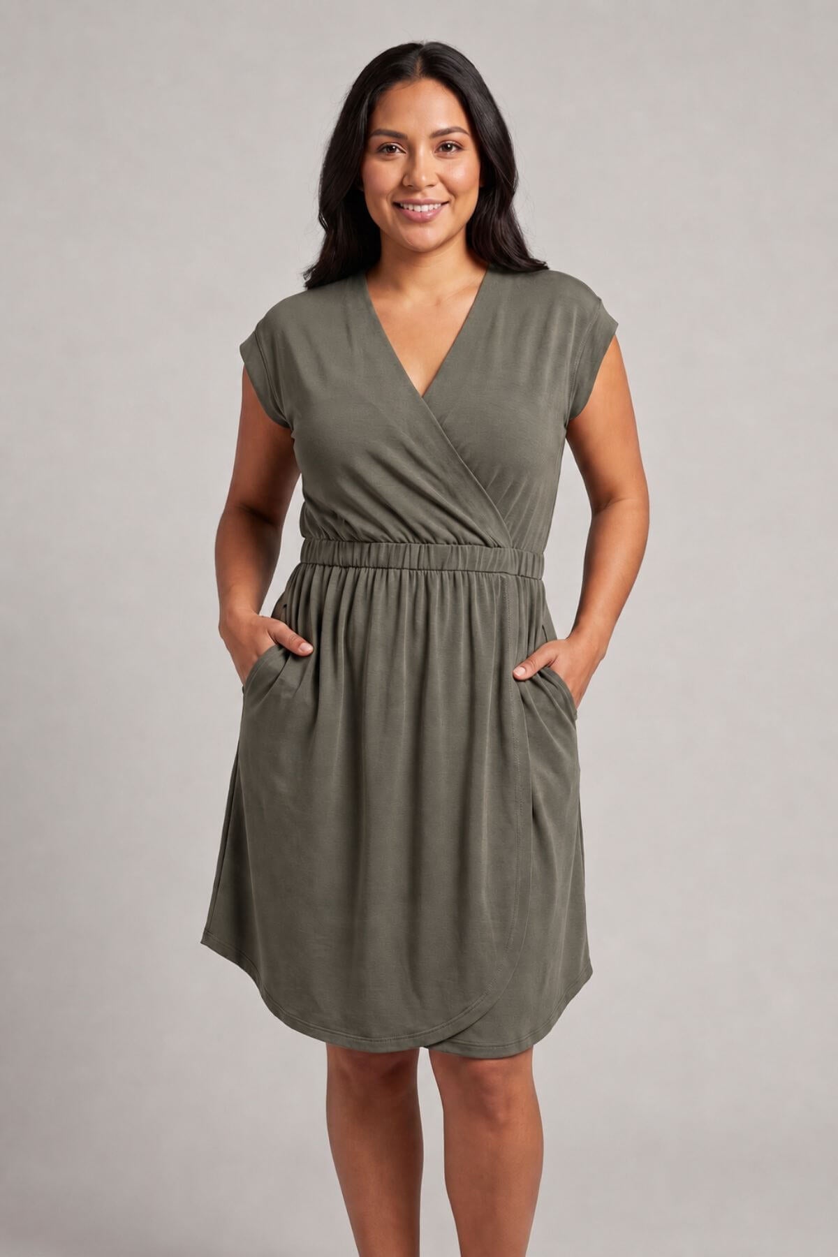 A woman modeling an olive green wrap-style dress with short cap sleeves, side pockets, and a gathered waist, photographed against a plain light gray studio background..