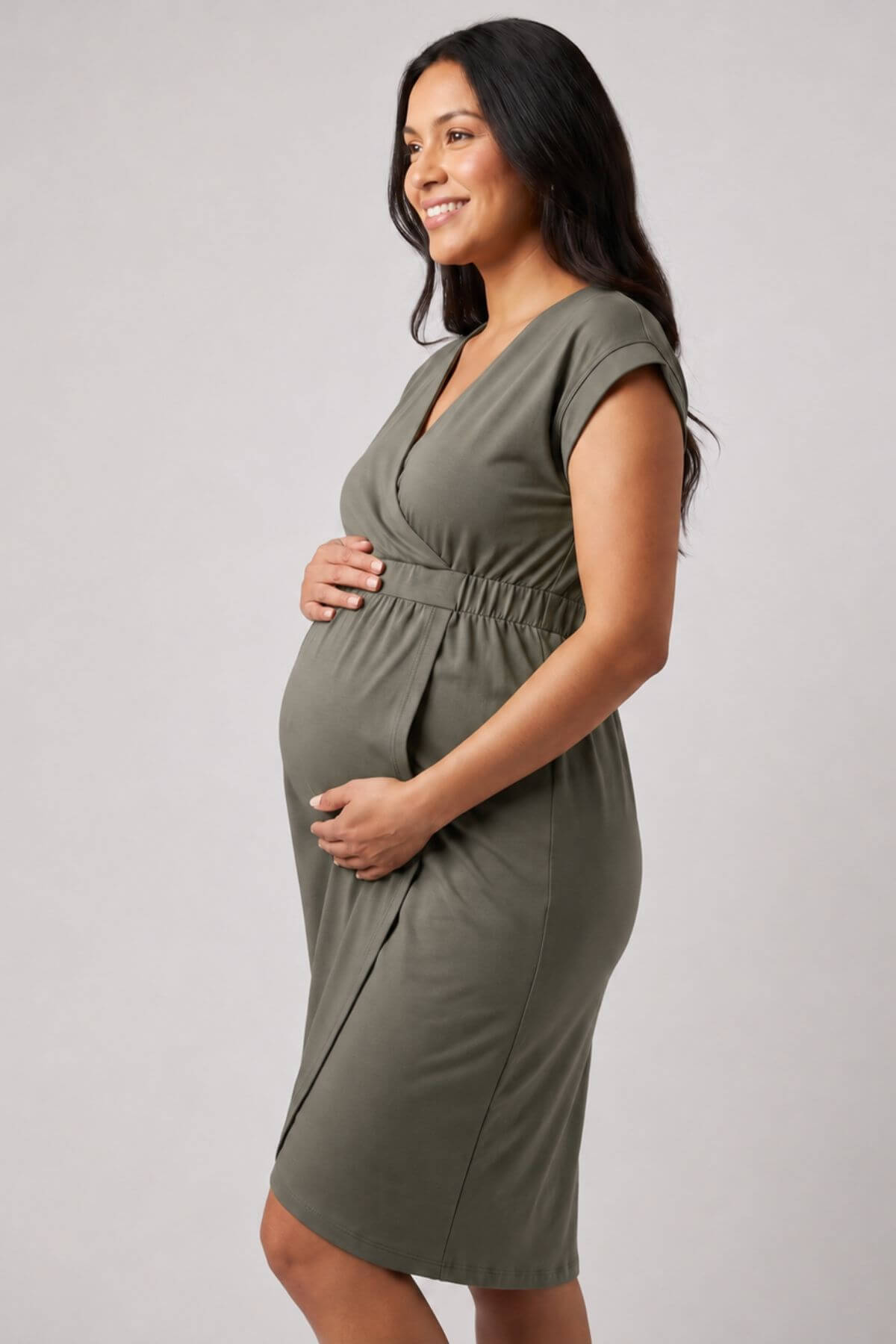 A pregnant woman wearing an olive green maternity dress, standing in profile against a light gray studio background.