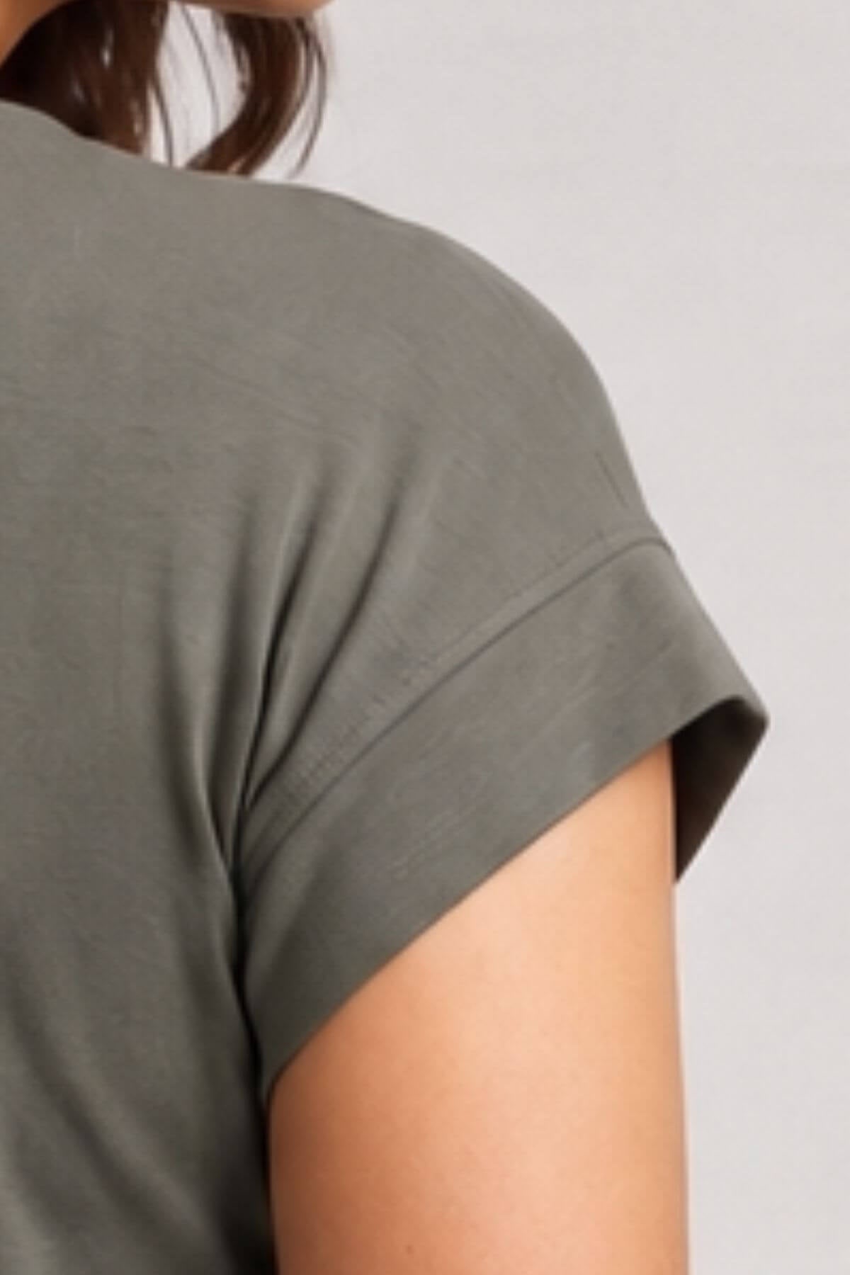 Close-up of a person wearing a short-sleeve crew-neck t-shirt in a muted olive green color, showing the side profile and sleeve cuff against a light gray background..