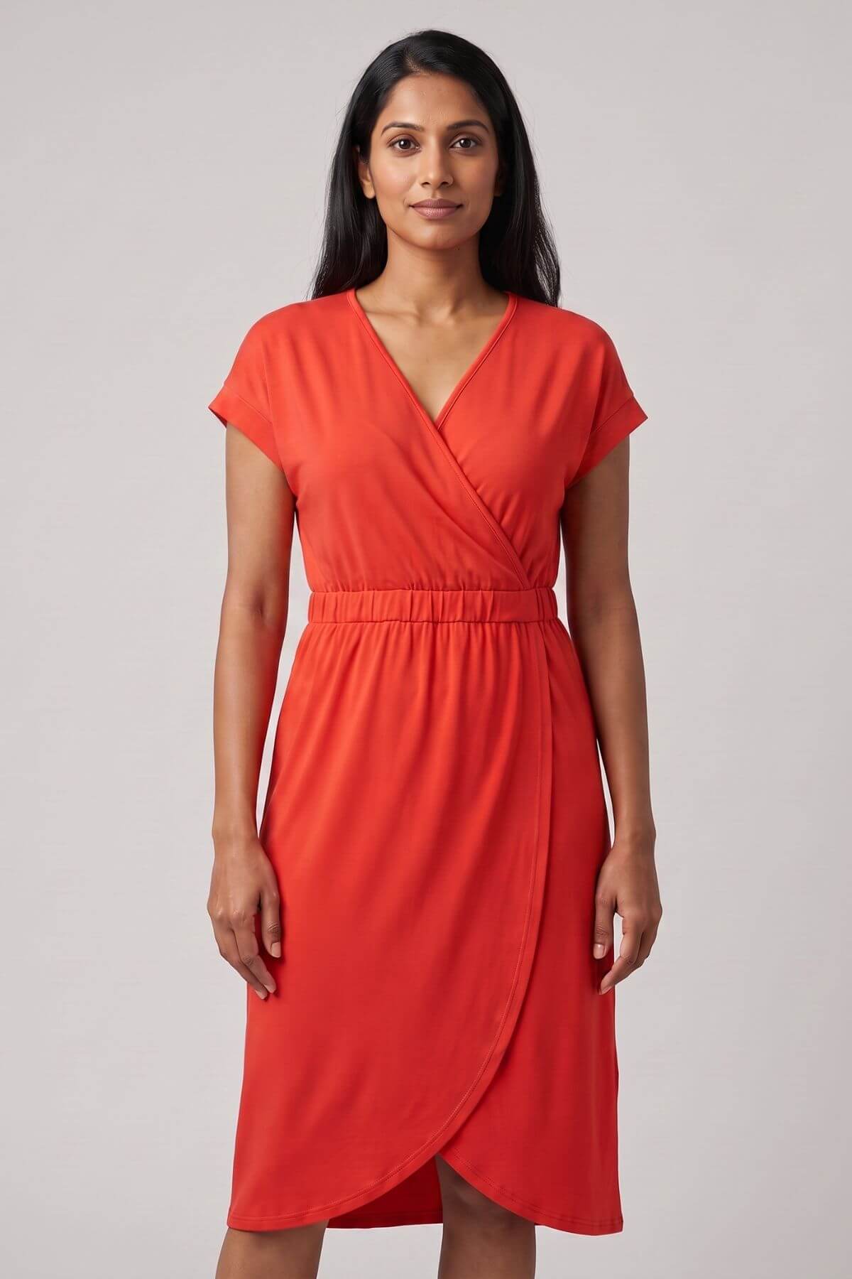 A woman modeling a bright coral-red wrap-style dress with short cap sleeves, a V-neckline, and a gathered waist, photographed against a plain light gray studio background.
