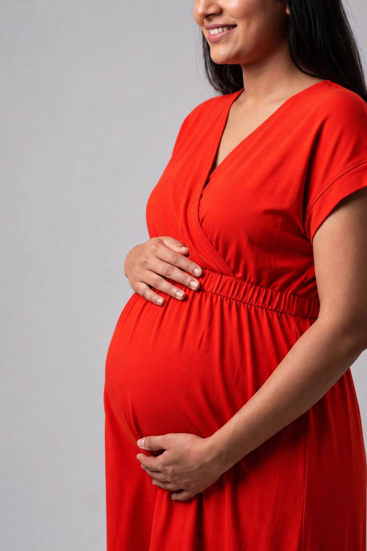 A pregnant woman wearing a vibrant red wrap-style maternity dress, standing against a neutral gray studio background.