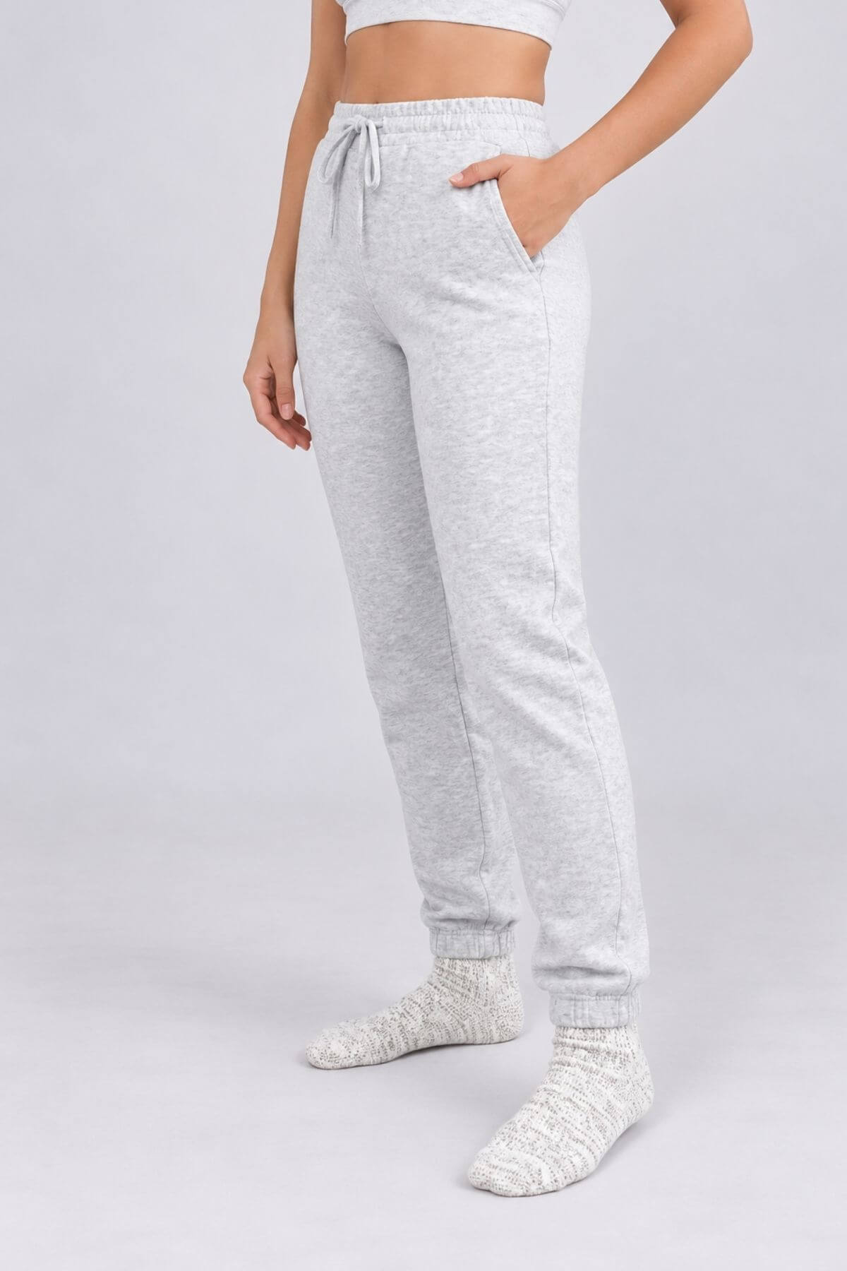 A person modeling light heather gray jogger sweatpants with a drawstring waist, side pockets, and ribbed ankle cuffs, paired with white socks against a light gray studio background..