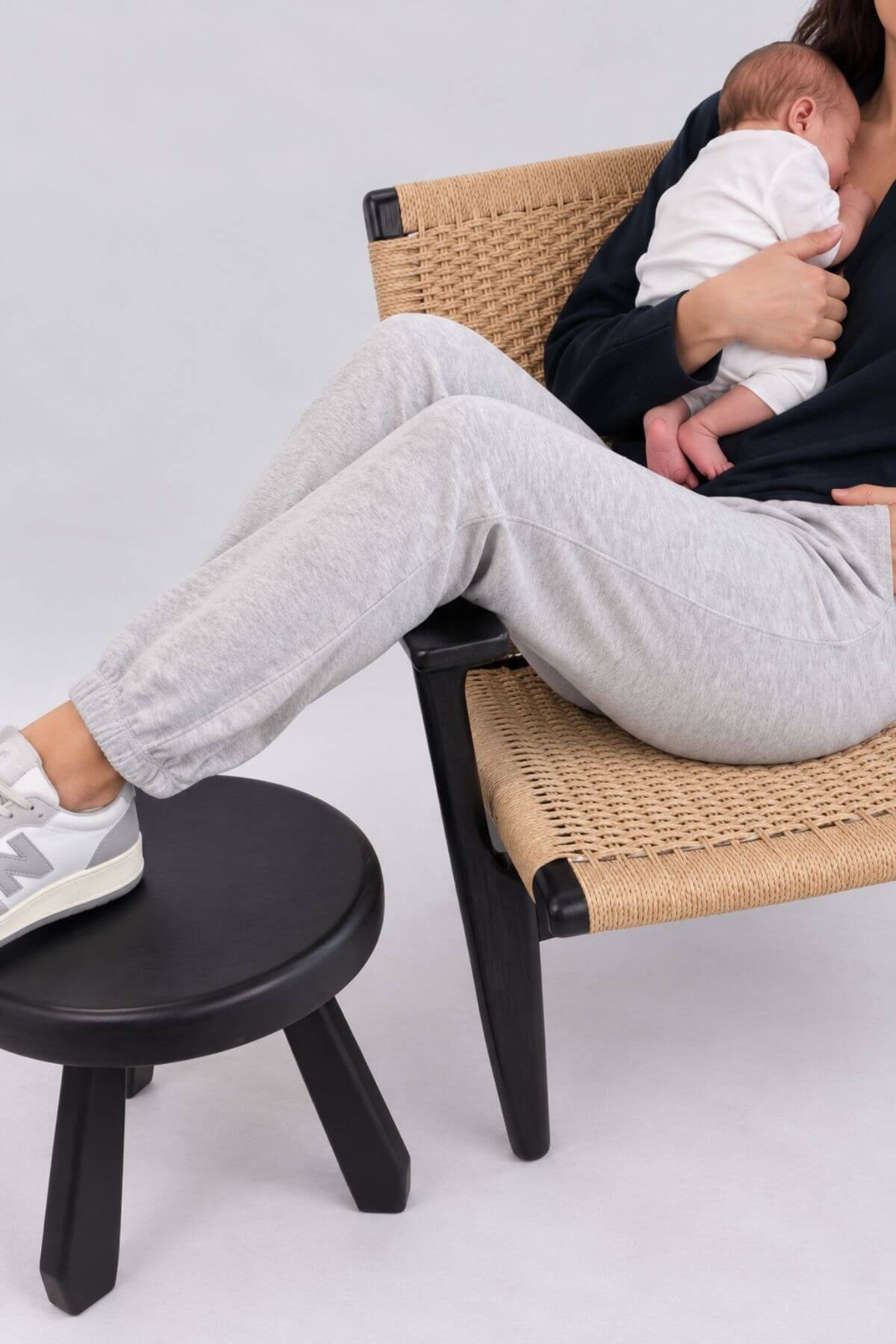 A person sitting on a woven chair with a baby in their arms, with one foot resting on a small black stool. The person is wearing light gray sweatpants, a dark top, and white New Balance sneakers.