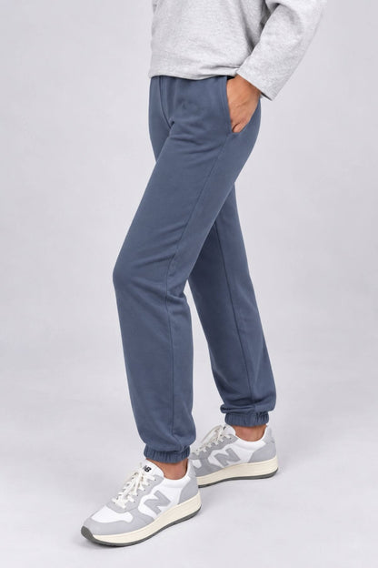 A person wearing slate blue jogger pants with elastic cuffs, paired with a light gray sweatshirt and gray New Balance sneakers, shown from the waist down against a light gray studio background.
