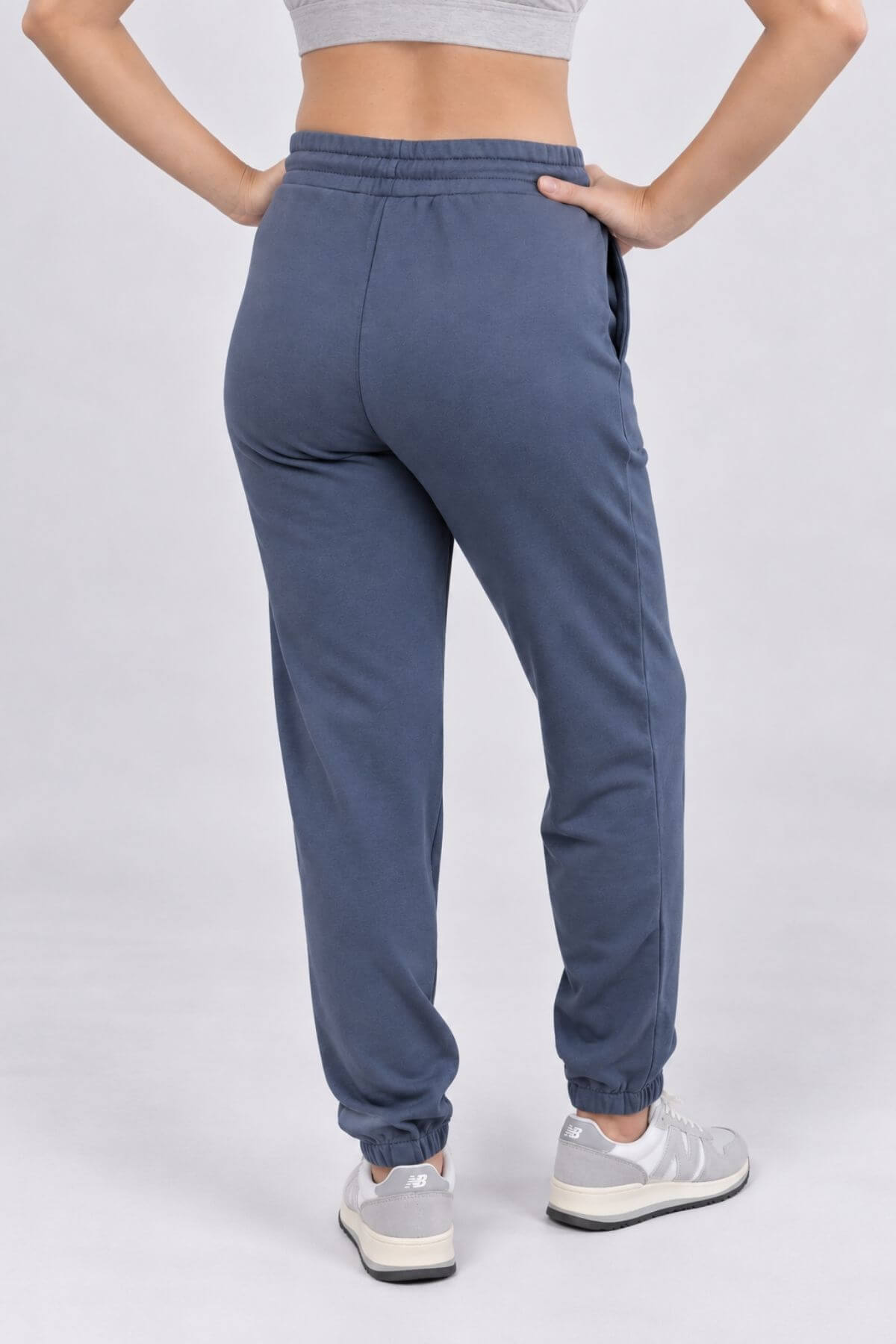 A person shown from the back wearing slate blue jogger sweatpants with elastic cuffs, paired with a gray sports bra and white sneakers, against a light gray studio background.