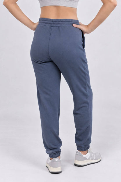 A person shown from the back wearing slate blue jogger sweatpants with elastic cuffs, paired with a gray sports bra and white sneakers, against a light gray studio background.